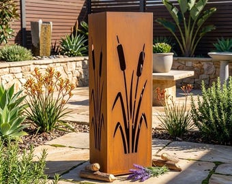 May include: A tall, rectangular, rust-colored metal garden sculpture featuring laser-cut silhouettes of cattail plants on its sides. The structure stands upright on a stone patio, with small pieces of wood and lavender sprigs placed at its base.