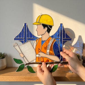 May include: Stained glass art piece depicting a construction worker in a yellow hard hat and orange safety vest, holding a blueprint. The background features a blue bridge design. The artwork is accented with green leaves and a silver chain.