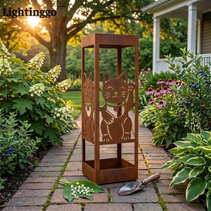 Puede incluir: Una linterna de jardín alta y rectangular de metal color óxido con diseños recortados de gatos de la suerte en sus lados. La estructura tiene un diseño de marco abierto con una base y una parte superior sólidas, con un acabado metálico oxidado.
