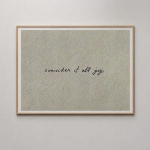 Op de afbeelding: Een rechthoekige kunstprint in een lichthouten lijst met een gestructureerde, beige, zandachtige achtergrond. Gecentreerd op de print staat de tekst "consider it all joy" geschreven in een zwart, cursief lettertype met kleine letters.
