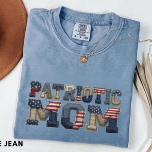Puede incluir: Una camiseta Comfort Colors azul claro con las palabras "PATRIOTIC MOM" bordadas en rojo, blanco y azul. Un collar dorado con un colgante descansa sobre la camiseta. La camiseta está doblada y tiene una etiqueta pequeña.