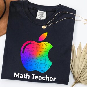 Puede incluir: Una camiseta azul marino con un diseño de manzana colorido lleno de ecuaciones matemáticas. La manzana es de colores arcoíris. Las palabras "Math Teacher" están impresas en blanco en la parte inferior de la camiseta.