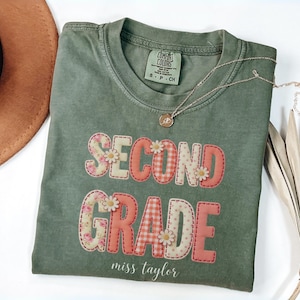 Puede incluir: Una camiseta de manga corta color verde oliva con el texto "SECOND GRADE" en estilo patchwork con patrones florales y de cuadros vichy, acentuado con pequeñas margaritas blancas. El nombre "miss taylor" está impreso en letra cursiva blanca.