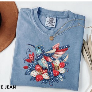 Puede incluir: Camiseta azul vaquero con un diseño de colibrí detallado. El colibrí tiene detalles en azul, rojo y blanco. La camiseta también tiene el texto "BLUE JEAN" en la parte inferior.
