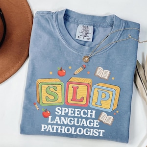 Puede incluir: Una camiseta azul con un gráfico de tres bloques de madera con letras que forman SLP, rodeados de ilustraciones de manzanas, lápices y libros abiertos. Debajo de los bloques, el texto SPEECH LANGUAGE PATHOLOGIST aparece en letras blancas.