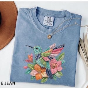 Puede incluir: Una camiseta azul vaquero con un diseño de colibrí colorido con alas y flores de retazos. La camiseta tiene cuello redondo y mangas cortas. Un collar de oro con un colgante está drapeado sobre la camiseta. El texto "BLUE JEAN" está en la parte inferior.