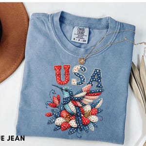 Puede incluir: Una camiseta azul vaquera con la palabra "USA" en letras de patchwork rojas, blancas y azules. Un colibrí con alas patrióticas y detalles florales está bordado debajo del texto. Un collar dorado está sobre la camiseta.