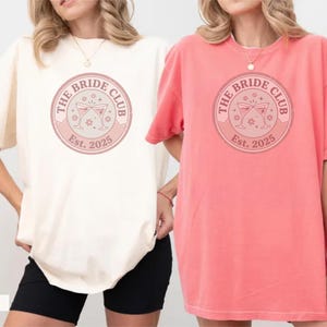 Puede incluir: Dos camisetas holgadas, una crema y otra coral, cada una con un diseño circular. El diseño dice "The Bride Club" con el año "Est. 2025" e incluye ilustraciones de copas de cóctel. Las camisetas son informales y cómodas.