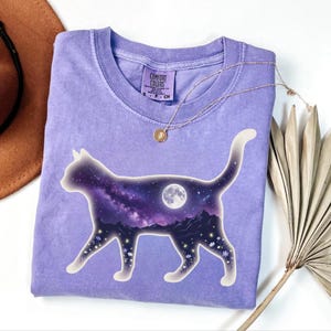 Puede incluir: Una camiseta de manga corta de color morado claro con un gráfico de la silueta de un gato relleno con una escena de cielo nocturno, que incluye una luna llena, una nebulosa morada, picos de montañas y pequeñas flores blancas.