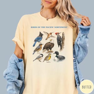 Puede incluir: Camiseta amarillo claro con una ilustración detallada de varias aves, incluyendo un arrendajo azul, un colibrí y un águila calva. El texto "BIRDS OF THE PACIFIC NORTHWEST" está impreso encima de las aves. La camiseta tiene mangas cortas y un corte relajado.