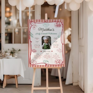 May include: A wooden easel displays a personalized sign for "Mattheo's Holy Cannoli Milestones." The sign features a photo of a baby in a chef's hat, surrounded by red and white decorative elements. The sign lists milestones and things the baby loves.