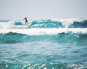 Puede incluir: Un surfista surfea una ola en el océano. La imagen captura la silueta del surfista contra una gran ola turquesa. El agua es azul oscuro y el cielo está despejado.