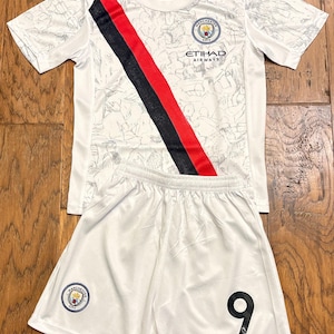 May include: A white Manchester City football kit comprising a shirt and shorts. The shirt features a diagonal black and red stripe, a faint grey graphic pattern, the Manchester City crest, and Etihad Airways text. The shorts include the team crest and number 9.