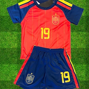 May include: A red and blue soccer jersey and shorts set featuring the number 19 in yellow on both pieces. The jersey has yellow stripes on the shoulders and the Spanish national crest on the chest. The blue shorts include the crest and number 19.