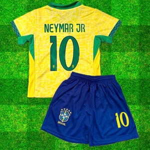 Puede incluir: Un conjunto de camiseta y pantalón corto de fútbol amarillo y verde. La camiseta lleva el nombre "NEYMAR JR" y el número "10" en la espalda. El pantalón corto azul también muestra el número "10" y un logo del equipo brasileño. Sobre un fondo verde.