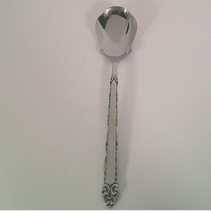 May include: A silver-colored serving spoon with a decorative handle and a petal-shaped bowl. The handle features an ornate design, and the spoon is set against a white background. This utensil is likely for serving food.