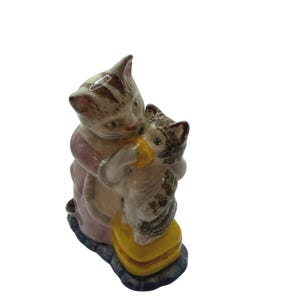 May include: A ceramic figurine depicting two cats embracing. The larger cat, in a pink and cream-colored outfit, hugs a smaller cat. The cats are primarily gray and brown with striped patterns. They are sitting on a yellow cushion.