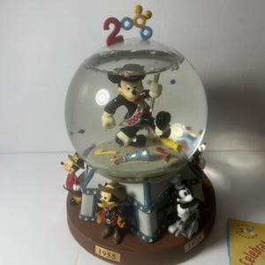Op de afbeelding: Een Disney sneeuwbol met Mickey Mouse in een zwart en rood uniform in een glazen bol. De basis heeft Mickey Mouse figuren in verschillende outfits, met de nummers "1955" en "1928" op de basis. De bovenkant van de bol heeft het nummer "2008".