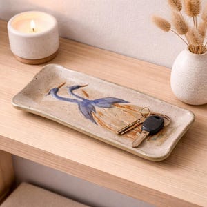 May include: A rectangular ceramic tray with a hand-painted design of two blue herons. The tray holds a set of keys. A lit candle and a small vase with dried flowers are in the background. The tray is approximately 25 cm long.