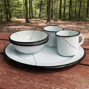 May include: A white speckled enamelware camping set with black trim. The set includes a large plate, two bowls, and two mugs. Each piece has a black dotted design. The set is on a wooden table in a forest setting.