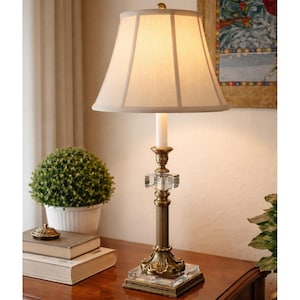 Puede incluir: Lámpara de mesa decorativa con pantalla beige y base de latón. La lámpara presenta un detalle de cristal y una base cuadrada. Se encuentra sobre una superficie de madera, junto a una pequeña planta en maceta y libros. Altura: aprox. 60 cm.