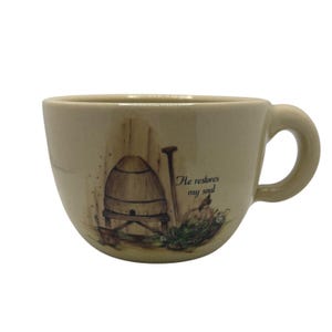 May include: Cream-colored ceramic teacup with a rounded shape and a handle. The cup features a pastoral scene with a beehive, a pumpkin, and the text "He restores my soul."