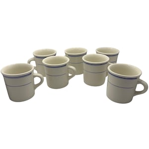 Może przedstawiać: Zestaw siedmiu kremowych ceramicznych kubków z niebieskim paskiem. Każdy kubek ma zakrzywiony uchwyt i prosty, klasyczny design. Kubki są ułożone w grupie, odpowiednie do kuchni lub jadalni.