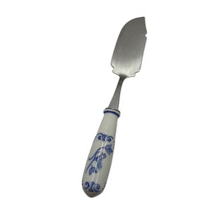 Puede incluir: Un cuchillo de queso de acero inoxidable con un mango de porcelana blanca. El mango está decorado con motivos florales azules y un borde azul. El cuchillo tiene una hoja curva con borde dentado.