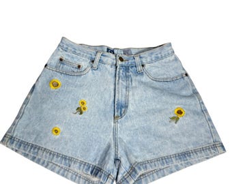 Puede incluir: Pantalones cortos de mezclilla azul claro con efecto lavado ácido, diseño de cintura alta, cierre de botón y cremallera. Los pantalones cortos están decorados con pequeños motivos bordados de girasoles amarillos con tallos verdes.
