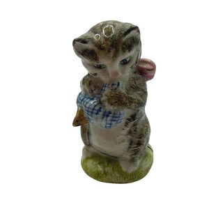 May include: A ceramic figurine of a kitten, with gray and brown tabby markings, holding a blue and white checkered bag. The kitten has green eyes and a pink bow. The figurine is standing on a patch of green grass.