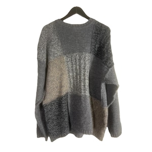 Op de afbeelding: Een patchwork trui in tinten grijs, beige en zwart hangt aan een zwarte hanger. De trui heeft een ronde hals en een mix van getextureerde gebreide panelen. Het ontwerp is een mix van effen en gespikkelde garens.