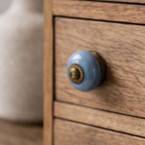 May include: Close-up of a wooden drawer with a light blue ceramic knob. The knob has a brass base and a detailed design. The wood grain of the drawer is visible, showing a natural, warm tone. The knob adds a decorative touch to the furniture.