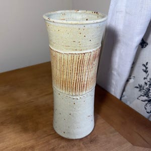 May include: A tall, cylindrical ceramic vase with a speckled off-white glaze. The upper and lower sections feature a smooth finish, while the central band is decorated with vertical, incised lines in a warm, earthy brown tone.