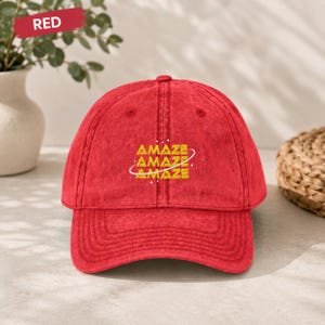 May include: A red baseball cap with a distressed, washed-out texture. The front features the word "AMAZE" embroidered in yellow three times, stacked vertically, with a thin white circular line orbiting the text and small white star accents.