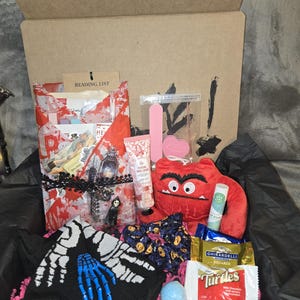Pode incluir: Uma caixa de presente cheia de vários itens, incluindo um monstro de pelúcia vermelho, meias pretas com desenhos de mãos de esqueleto, creme para as mãos e uma lista de leitura. A caixa também contém bombas de banho, chocolate e uma pequena bolsa de artigos de beleza.