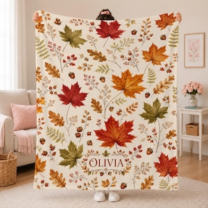 May include: A cream-colored throw blanket featuring a repeating autumn pattern of red, orange, and green maple leaves, oak leaves, ferns, and brown acorns. The name OLIVIA is printed in brown serif lettering within a decorative leaf-shaped frame.