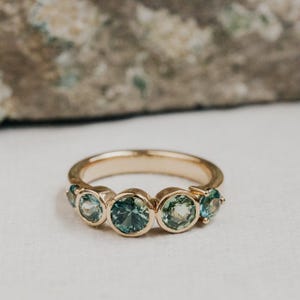 Könnte beinhalten: Ein Goldring mit fünf rund geschliffenen, blaugrünen Edelsteinen in Zargenfassung. Die Steine variieren in der Größe, wobei der größte Edelstein in der Mitte sitzt und von zwei kleineren Steinen auf jeder Seite des polierten Goldbandes flankiert wird.