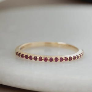 Könnte beinhalten: Ein goldener Bandring mit einer Reihe kleiner, runder, roter Edelsteine. Der Ring wird auf einer weißen Oberfläche präsentiert, wobei die weiche Beleuchtung die Details hervorhebt. Der Ring ist ein zartes Schmuckstück.