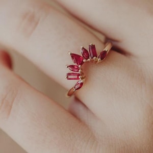 Könnte beinhalten: Ein goldener Ring mit einem halbkreisförmigen Design aus leuchtend roten Edelsteinen. Der Ring wird an einem Finger getragen und unterstreicht die komplizierten Details des Schmucks. Die Edelsteine sind rechteckig geschliffen.