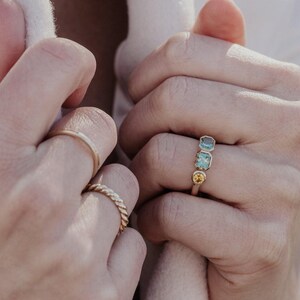 Puede incluir: Primer plano de manos con anillos de oro. Una mano muestra un anillo con tres piedras preciosas: dos azules y una amarilla. Otra mano tiene una banda de oro simple y un anillo de oro trenzado.