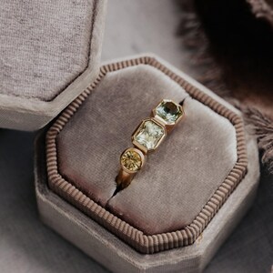 Puede incluir: Un anillo de oro con tres piedras preciosas: una piedra amarilla redonda, una piedra verde claro rectangular y una piedra verde claro hexagonal. El anillo se muestra dentro de un joyero de terciopelo gris.