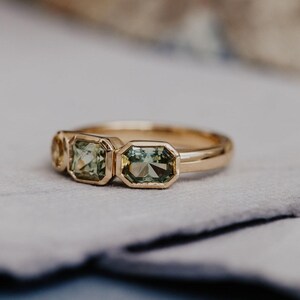 Puede incluir: Un anillo de oro con tres piedras preciosas de talla esmeralda en tonos verdes y amarillos. El anillo tiene una banda de oro pulido y engastes de bisel. El anillo se muestra sobre una superficie gris claro.