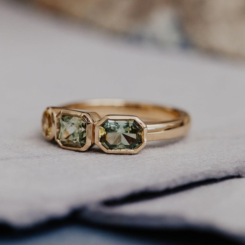 Puede incluir: Un anillo de oro con tres piedras preciosas de talla esmeralda en tonos verdes y amarillos. El anillo tiene una banda de oro pulido y engastes de bisel. El anillo se muestra sobre una superficie gris claro.