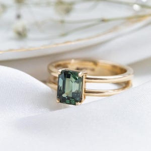 Könnte beinhalten: Ein goldener Ring mit doppeltem Banddesign, mit einem rechteckigen, smaragdgeschliffenen grünen Edelstein. Der Ring ist vor einem weißen Stoffhintergrund mit weicher Beleuchtung platziert.