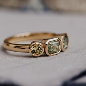 Puede incluir: Un anillo de oro con tres piedras preciosas. El anillo tiene una gema amarilla redonda, una gema verde claro de corte cuadrado y una gema verde claro. El anillo est&aacute; engastado en una banda de oro.