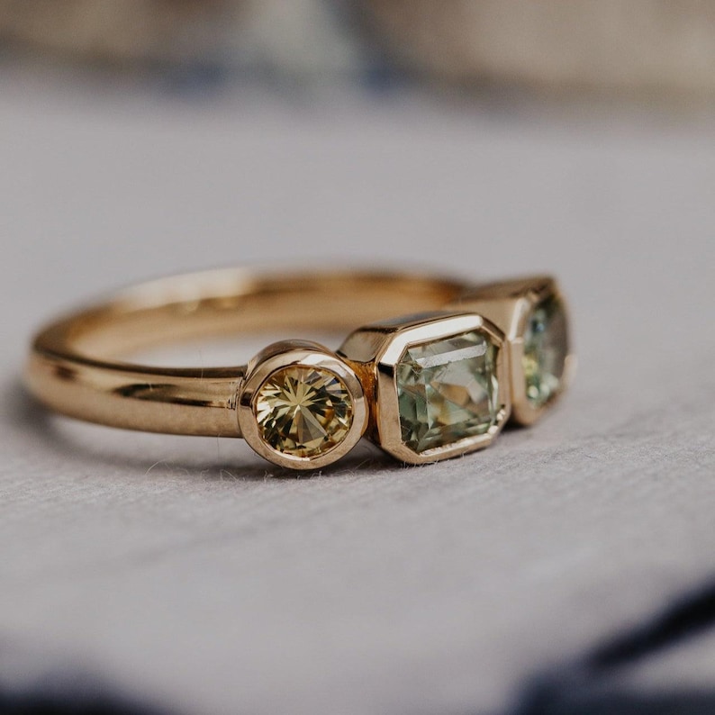 Puede incluir: Un anillo de oro con tres piedras preciosas. El anillo tiene una gema amarilla redonda, una gema verde claro de corte cuadrado y una gema verde claro. El anillo est&aacute; engastado en una banda de oro.
