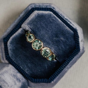 Peut inclure: Une bague en or ornée de cinq pierres précieuses rondes vert clair, disposées en ligne. La bague est présentée dans un écrin de velours bleu. Les pierres sont facettées et captent la lumière.