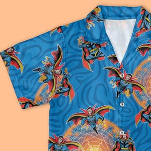 May include: A short-sleeved shirt with a blue background featuring a repeating pattern of a superhero. The superhero is depicted in various action poses, wearing a red cape and blue outfit, with orange and yellow energy circles.