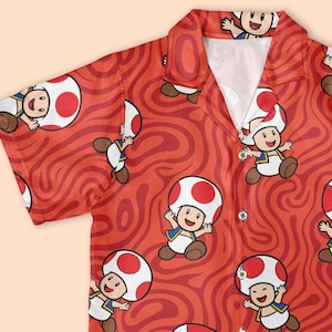 May include: Red button-down shirt featuring a repeating pattern of a cartoon character with a mushroom-shaped head. The shirt has short sleeves and a white collar. The background is a swirling red design.