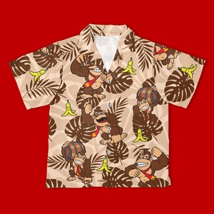 May include: A short-sleeve button-down shirt in a light tan color featuring an all-over print of brown gorillas wearing red ties, yellow bananas, and dark brown tropical palm leaves. The shirt has a classic collar and a white inner neckline.